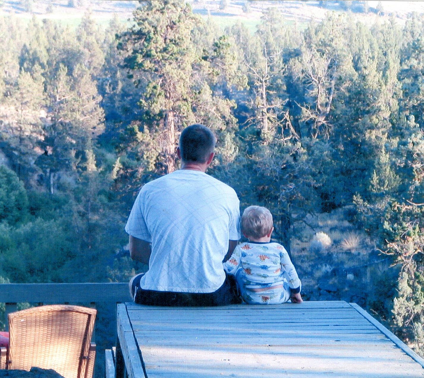 Father and son sitting together on a deck overlooking a forested landscape